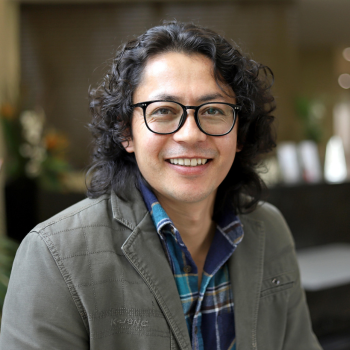 Smiling man with curly hair and glasses wearing a jacket and checkered shirt indoors.